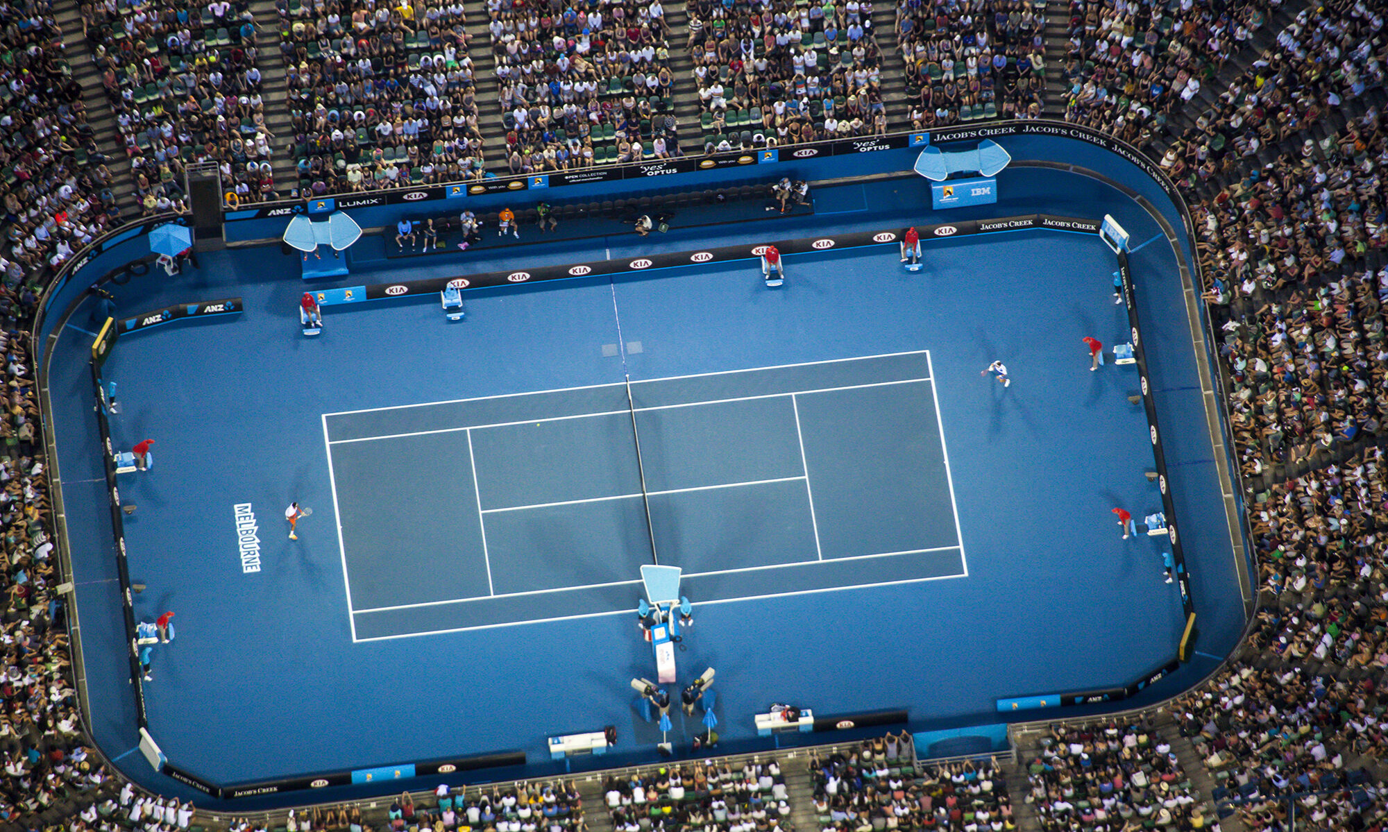 Aerial view of a professional tennis match in a large stadium. The blue tennis court is surrounded by a full crowd of spectators. Players and officials are visible on the court.