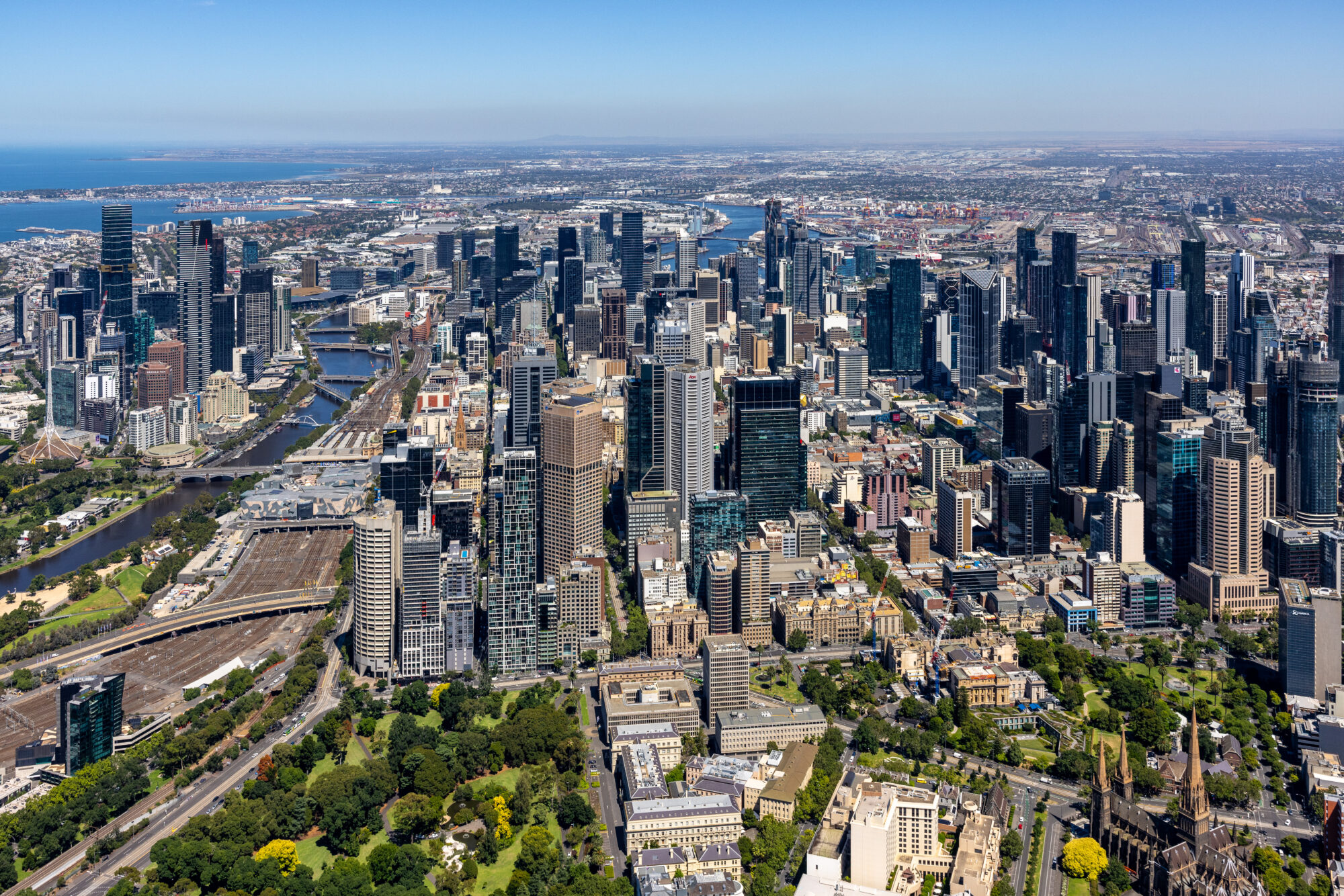 Aerial view of the Melbourne CBD skyline, featuring the Yarra River, Flinders Street Station tracks, and Port Phillip Bay