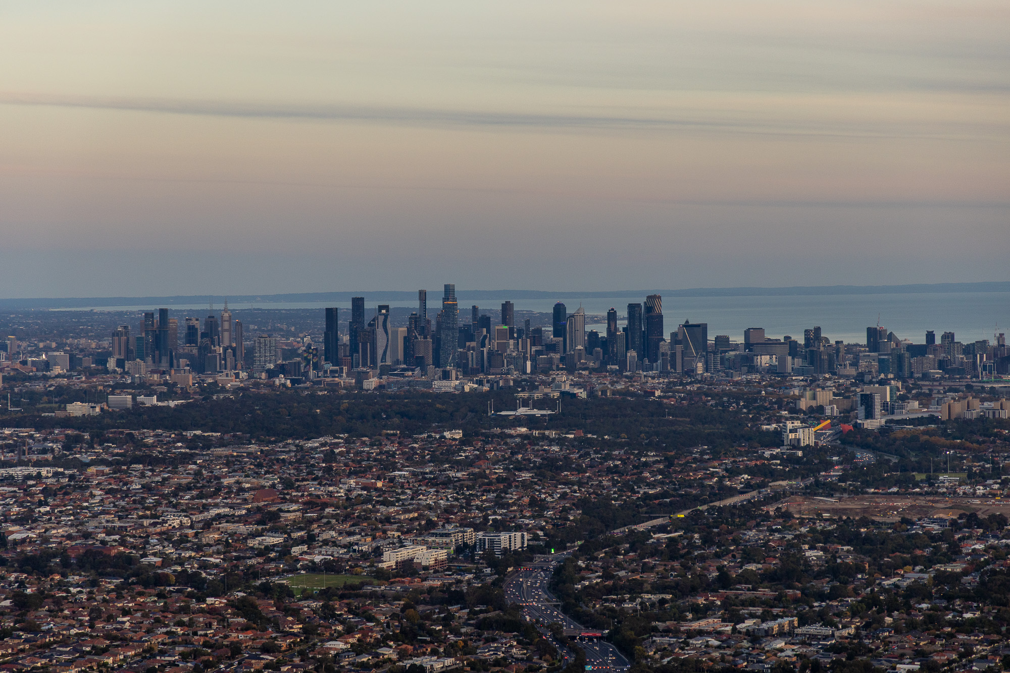 Helicopter aerial photography of Melbourne CBD skyline at dusk, with Port Phillip Bay in the background and CityLink in the foreground